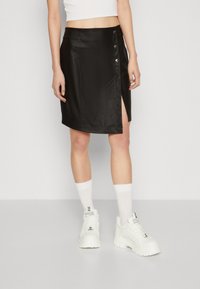 Zwarte imitatielederen rok met een zijsplit, voorzien van drukknoopdetail. Gladde textuur, aansluitend ontwerp en lengte tot halverwege de dijen.