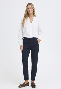 Weiße Bluse mit langen Ärmeln und Dekolleté-Detail, kombiniert mit marineblauen, taillierten Hosen und braunen Loafern. Glattes Material und maßgeschneiderte Passform.