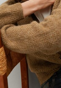 Pull marron tricoté avec une texture épaisse et des poignets côtelés, porté par-dessus un haut blanc, avec une chaise en bois partiellement visible en arrière-plan.