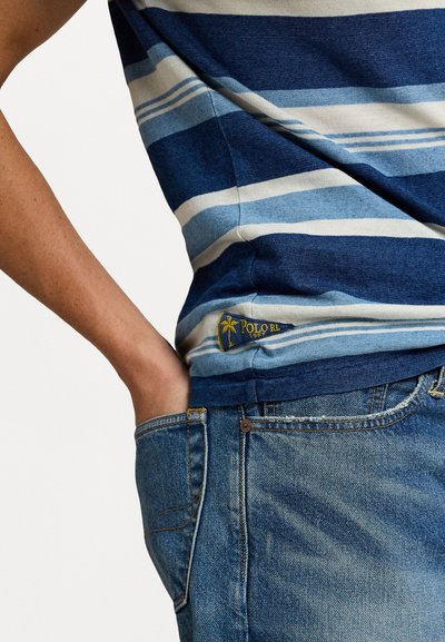 Gestreepte katoenen t-shirt in tinten blauw en crème, met een klein logo label op de zoom. Draagt een spijkerbroek.