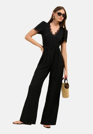 Kvinde iført en sort jumpsuit med blondekant, sorte solbriller, der holder en flettet håndtaske og iført sorte sandaler mod en ensfarvet baggrund.
