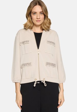 Frau trägt eine beige Jacke mit Reißverschluss, vier Klappentaschen und Kordelzug am Saum, kombiniert mit schwarzen Hosen, stehend vor weißem Hintergrund.