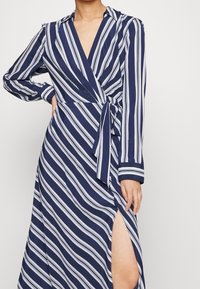 Tmavě modré zavinovací šaty s bílými vertikálními pruhy, dlouhými rukávy, hlubokým výstřihem do V, zavázáním v pase a bočním rozparkem pro zvýraznění detailu.