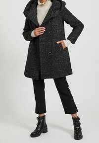 Svart texturerad ullkappa med stor huva, knappstängning och sidofickor, kombinerad med svarta byxor och snörda svarta stövlar.