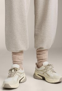Sneaker bianche e beige in materiale mesh e sintetico, con suole testurizzate e calzini a coste rosa chiaro sotto pantaloni grigi.