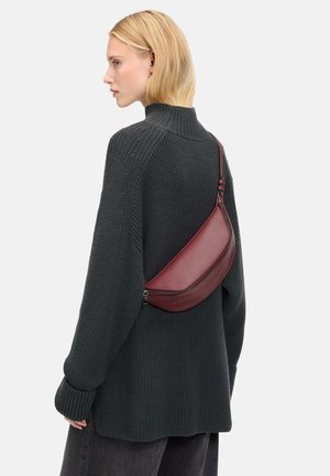 Borsa a tracolla in pelle bordeaux con forma curvata e chiusura con zip, indossata sopra un maglione di lana scuro, accentuato da una texture a coste.