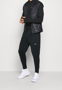 Zwarte mouwloze puffer vest over een zwart langemouwen top, gecombineerd met een zwarte tapered broek met een Nike-logo en witte sportschoenen.