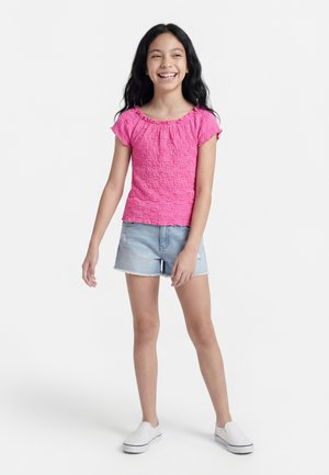 Κορίτσι που χαμογελά με μακριά μαύρα μαλλιά, ντυμένο με ροζ μπλούζα κοντών μανικιών με υφή, ανοιχτό μπλε σορτς από τζιν και λευκά παπούτσια χωρίς κορδόνια, όρθια.