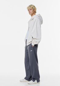 Lichtgrijze oversized hoodie met een ritssluiting aan de voorkant en trekkoorden, gecombineerd met donkergrijze joggingsbroek met zijzakken en een logo-detail.