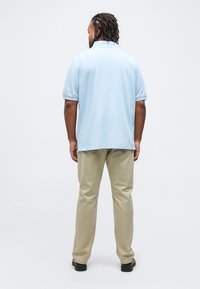 Άνδρας που στέκεται με την πλάτη γυρισμένη, φοράει ένα φωτεινό γαλάζιο κοντομάνικο πόλο, μπεζ παντελόνι και μαύρα παπούτσια, μπροστά σε μια απλή λευκή επιφάνεια.