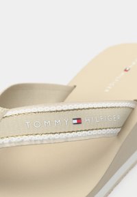 Beige flip-flop med en hvit og gull stropp med "TOMMY HILFIGER" merkevare og en flerfarget logo tag. Myk teksturert såle.