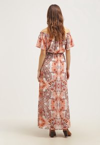 Kobieta z długimi, falującymi włosami nosi sukienkę bez ramiączek, sięgającą do podłogi, w pomarańczowo-brązowe kwiatowe wzory paisley, stojąc tyłem.