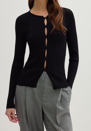 Femme portant un cardigan côtelé noir avec sept boutons sur le devant et un pantalon plissé gris à fines rayures, mains dans les poches.