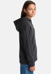 Tienermeisje met lang bruin haar, gekleed in een zwarte hoodie met rits en blauwe spijkerbroek, staand zijwaarts met een lichte glimlach.