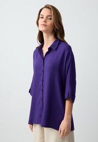 Chemise violette à boutons avec col, manches trois-quarts et texture douce. Présente une coupe décontractée et de légentes fentes sur les côtés à l'ourlet.