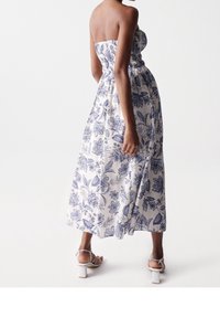 Olkaimeton valkoinen maksimekko sinisellä kukkakuviolla, jossa on kevyesti kuminauhalla kiristetty yläosa ja laskeutuva helma, yhdistetty valkoisiin lohkopohjaisiin sandaaleihin.