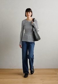 Gestreiftes Langarmhemd in Schwarz und Weiß, blaue Jeans mit ausgestelltem Bein, schwarze Schuhe und eine graue Tragetasche. Holzboden und schlichte Hintergrund.