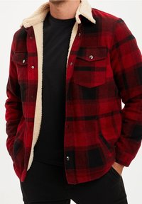 Man in een rood en zwart geruite jas met een crèmekleurige sherpa voering over een zwart shirt, met handen in de zakken.