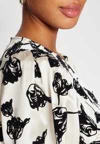 Impression florale noire sur un tissu blanc, avec une texture satinée lisse. Le design comprend un léger froncé au niveau de l'encolure.