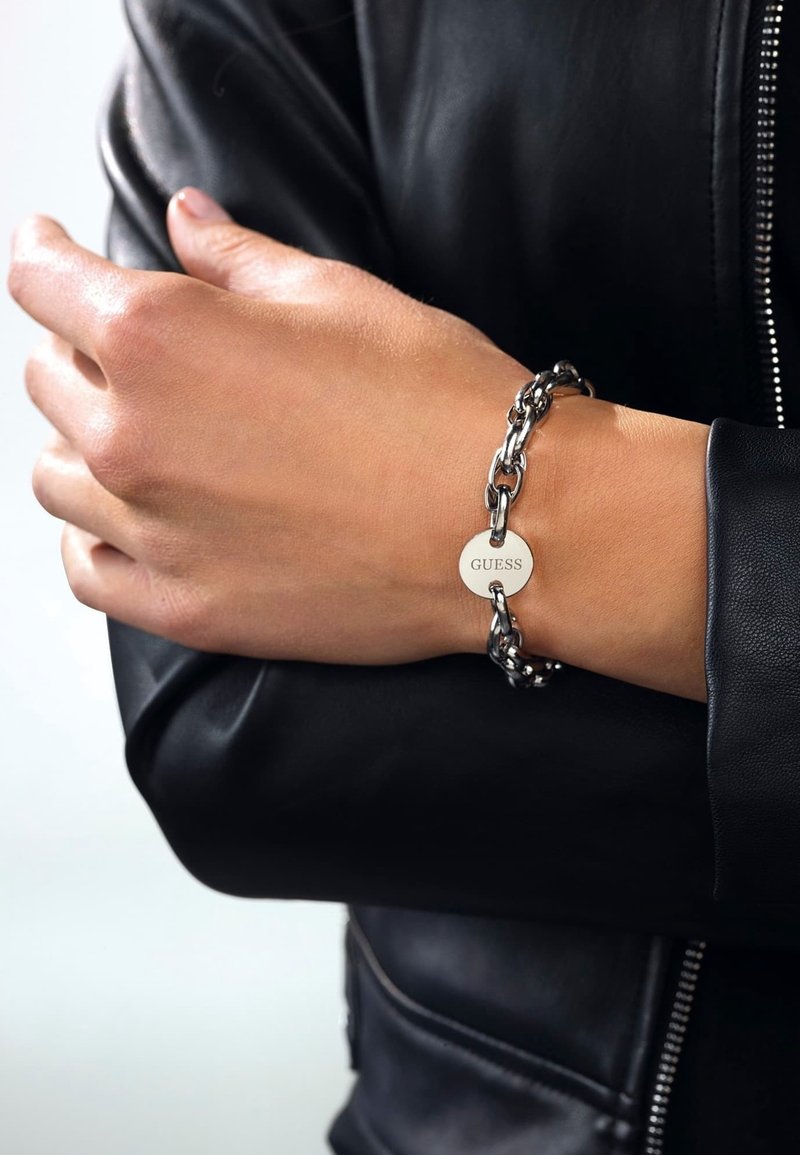 Gros plan d'une personne portant un bracelet chaîne en argent avec une étiquette ronde "GUESS" au poignet, vêtue d'une veste en cuir noire.