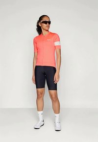 Radbekleidung bestehend aus einem leuchtend pinken kurzärmeligen Trikot mit weißen Akzenten und schwarzen Shorts. Getragen mit Sonnenbrille und weißen Radschuhen.