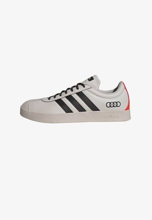 Bílá kožená teniska s černými pruhy a detaily, s gumovou podrážkou a malým červeným logem Audi na patě. Texturovaný povrch.