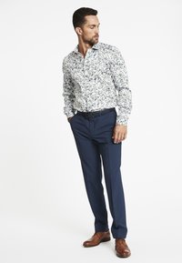 Langärmliges, floral gemustertes Hemd in Weiß mit blauen und grünen Mustern, kombiniert mit marineblauen, maßgeschneiderten Hosen und braunen Lederschuhen.