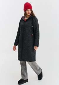 Žena v prošívaném černém kabátu po kolena, šedých džínách, černých bezklenkových botách a červené pletené čepici, stojící na jednobarevném bílém pozadí.