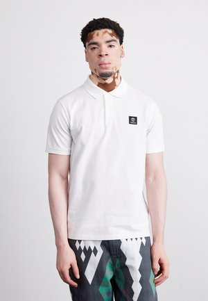 Polo shirt bianco in cotone con una toppa nera del logo, caratterizzato da un colletto classico e maniche corte, abbinato a pantaloni scuri a fantasia.