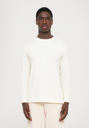 Man wearing plain long-sleeve off-white shirt and light beige pants, standing against a plain white background, facing forward.
