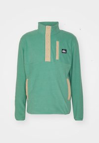 Pullover in pile verde con colletto alto, pattina con bottoni beige e due tasche laterali beige. Presenta un logo sulla parte superiore sinistra.