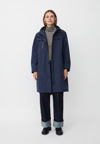 Cappotto impermeabile blu navy con cappuccio, due tasche frontali e chiusura con zip frontale. L'interno presenta un dolcevita a righe e pantaloni in denim.