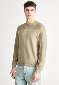 Beżowy sweter z dzianiny o fakturowanym wzorze, z okrągłym dekoltem i ściągaczami na rękawach. Na piersi naszyte logo. Połączony z jasnoniebieskimi dżinsami.