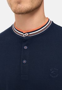 Marineblå polo skjorte med en knappet krave med orange og hvide striber. Den har tre blå knapper og et diskret logo på brystet.