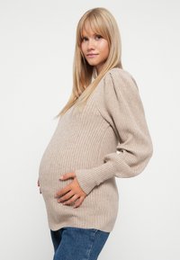 Beige ribbestrikket genser med lange, puffede ermer, båret over en gravid silhuett. Kombinert med blå jeans som har en avslappet passform.