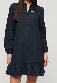Navy-blaues Hemdblusenkleid mit Rüsche am Saum, langen Ärmeln, Ausschnitten an der Brust und strukturierten Akzenten an den Ärmeln.