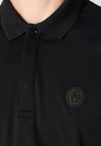 Black polo shirt made of cotton, featuring a ribbed collar and a single black button closure. Includes a round, black logo patch on the chest.