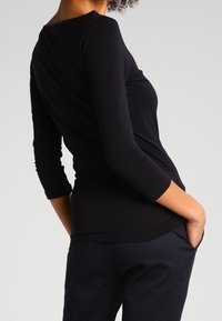 Maglietta nera a maniche lunghe con scollo rotondo e retro drappeggiato, abbinata a pantaloni scuri con texture, caratterizzati da un sottile motivo a zigzag.