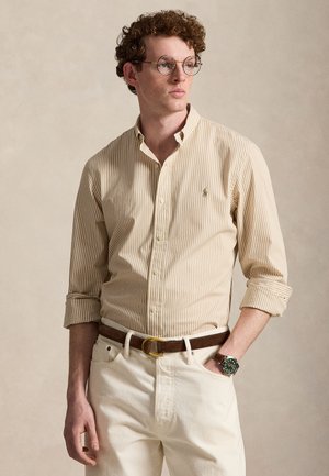 Young man with curly hair and glasses wearing a beige striped shirt, white pants, brown belt, and a green-faced wristwatch.