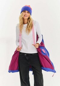 Barevná zateplená bunda ve fialové a růžové barvě s nastavitelnými pásky, s hladkým vnější povrchem a podšívkou uvnitř, nošená přes bílý top.