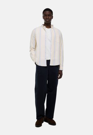 Homme portant une chemise à rayures jaunes claires boutonnée sur un henley blanc, un pantalon sombre et des chaussures marron, debout avec une main dans la poche.