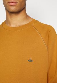 Sudadera amarilla mostaza con cuello redondo, textura suave y diseño de logo de bote bordado centrado en el pecho.