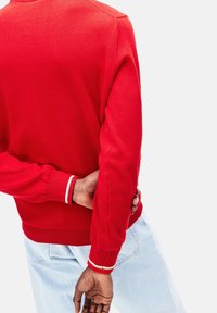 Rød sweater med ribbet halsudskæring og manchetter med kontrasterende hvide striber, lavet af strikket materiale, sammen med lyseblå jeans.