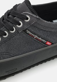 Sapatilhas de denim preto com sola de borracha, apresentando ilhós prateados, atacadores pretos e um rótulo de logotipo vermelho na lateral. Material superior texturizado.