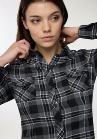 Camicia a quadri nera e grigia con chiusura a bottoni, tasche sul petto e colletto. Tessuto morbido con una finitura strutturata.