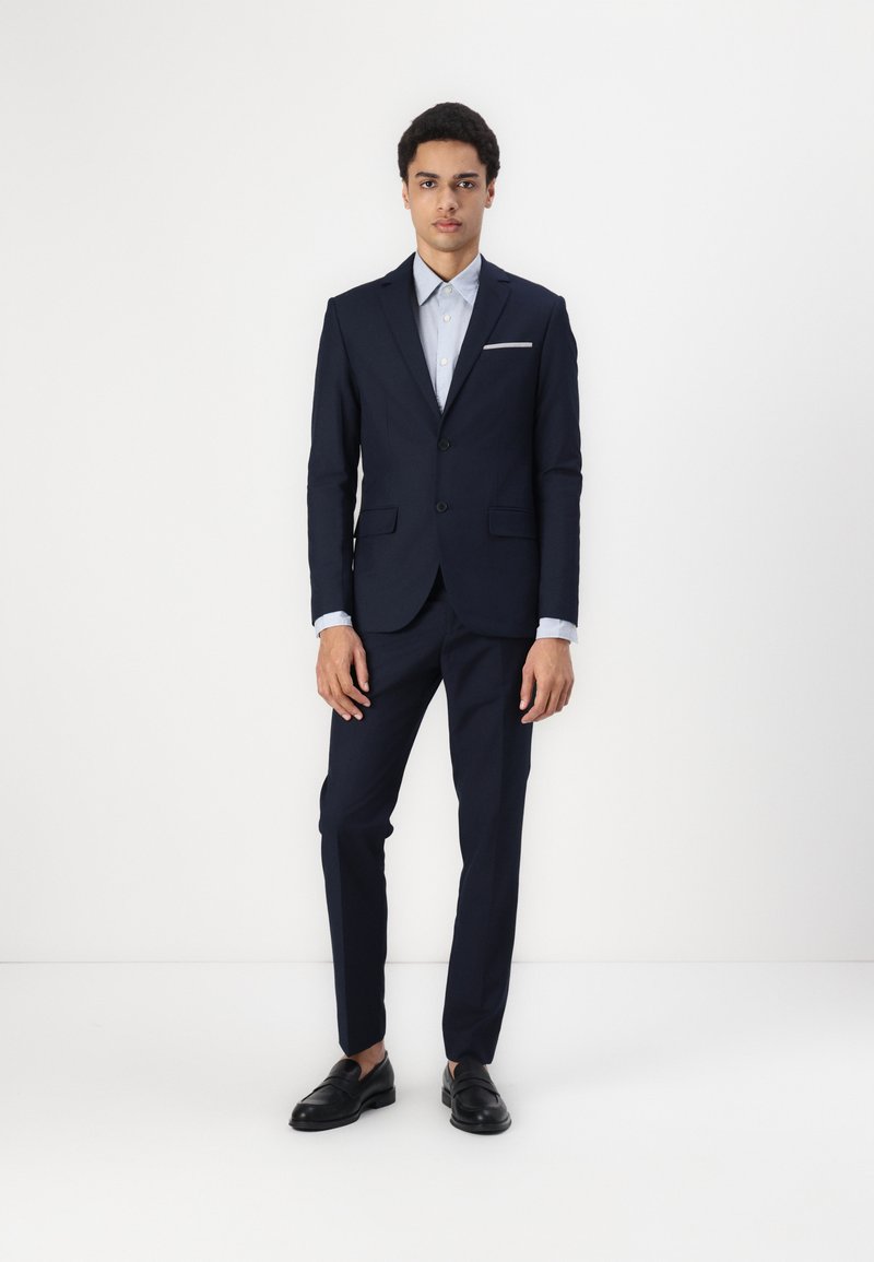 Navy suit with a tailored design, featuring a single-breasted jacket, notch lapels, and a light blue shirt. Black loafers complete the look.