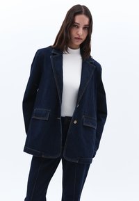 Tmavě modrý džínový blejzr s mírně oversize střihem, klopovými límci, dvěma předními kapsami a kontrastním oranžovým prošíváním. V kombinaci s odpovídajícími kalhotami.