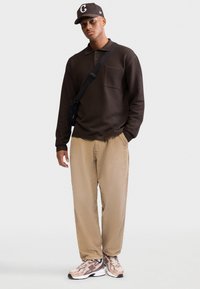 Polo marrón texturizado de manga larga con un bolsillo, combinado con pantalones de pana beige y zapatillas deportivas rosa claro. Bolsa bandolera negra y gorra.