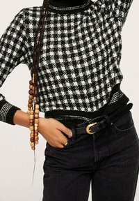 Pull en tricot noir et blanc à motifs à carreaux, avec un col rond et des poignets côtelés, associé à un jean noir et une ceinture noire avec une boucle en or.