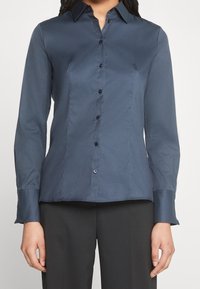 Camicia blu navy con bottoni, realizzata in tessuto liscio, caratterizzata da un colletto classico, maniche lunghe e bottoni scuri lungo la parte frontale.